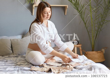 Cute gentle tender future mother wearing white clothing sitting in bed in light bedroom among baby's clothes choosing bodysuits for maternity hospital. 106537723