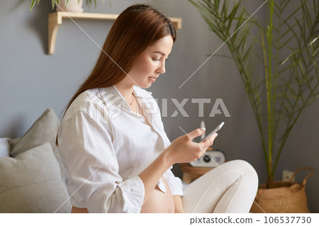 Profile portrait of adult pregnant woman with bare belly wearing white clothing sitting in bed in light bedroom, holding smart phone, browsing information about giving birth in internet. 106537730