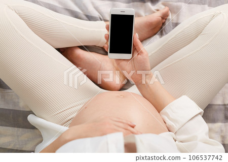 Unknown pregnant woman sitting in bed showing bare belly and smart phone with empty display, copy space for advertisement or promotional text. 106537742
