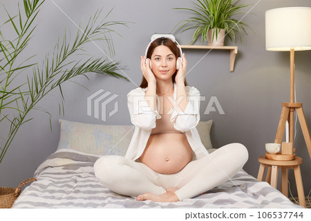 Pregnancy leisure. Calm relaxed pregnant brown haired caucasian woman sitting in bed with headphones listening to music spending leisure time at home. Pregnancy leisure. Calm relaxed pregnant brown haired caucasian woman sitting in bed with headphones listening to music spending leisure time at home. 106537744