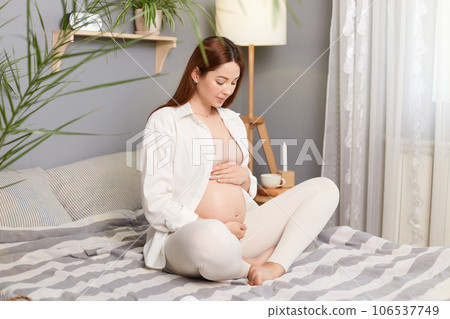 Attractive pregnant brown haired caucasian woman sitting in bed last months of pregnancy touching her tummy dreaming about first meeting with her child. 106537749