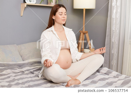 Meditating on maternity. Young caucasian pregnant woman practices yoga on bed at home in the morning Young beautiful pregnant woman sitting in lotus position good for fetus prenatal care. Meditating on maternity. Young caucasian pregnant woman practices yoga on bed at home in the morning Young beautiful pregnant woman sitting in lotus position good for fetus prenatal care. 106537754