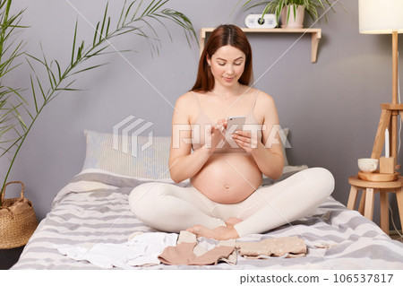 Pretty cute pregnant brown haired caucasian woman with bare belly sitting in bed surrounded with kids clothing using mobile phone browsing internet online shopping. 106537817