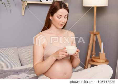 Caucasian expectant mother enjoying a cup of tea while sitting in bed enjoying moment of calmness and relaxation in her bedroom drinking hot beverage. Caucasian expectant mother enjoying a cup of tea while sitting in bed enjoying moment of calmness and relaxation in her bedroom drinking hot beverage. 106537820