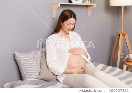 Cute charming pregnant brown haired caucasian woman wearing white shirt sitting in bed touching holding her belly relaxing resting on last months of pregnancy. Cute charming pregnant brown haired caucasian woman wearing white shirt sitting in bed touching holding her belly relaxing resting on last months of pregnancy. 106537821