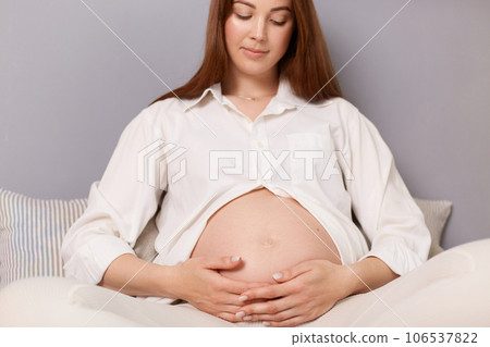 Pregnancy, motherhood, people and expectation. Calm pretty pregnant brown haired caucasian woman sitting in bed embracing her tummy resting at home. Pregnancy, motherhood, people and expectation. Calm pretty pregnant brown haired caucasian woman sitting in bed embracing her tummy resting at home. 106537822