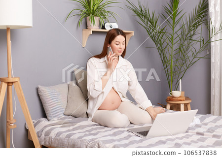 Busy pregnant brown haired caucasian woman sitting in bed working on laptop and talking on smart phone, combines maternity leave on last months of pregnancy and online work. Busy pregnant brown haired caucasian woman sitting in bed working on laptop and talking on smart phone, combines maternity leave on last months of pregnancy and online work. 106537838