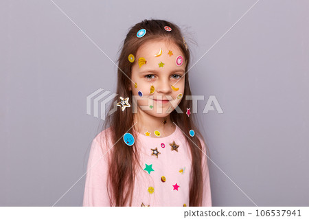 Positive cute little girl covered colorful kid's stickers standing isolated over gray background looking at camera with calm expression and charming smile. Positive cute little girl covered colorful kid's stickers standing isolated over gray background looking at camera with calm expression and charming smile. 106537941