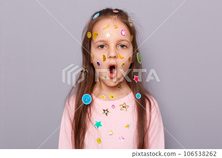 Surprised shocked little girl covered colorful kid's stickers standing isolated over gray background looking at camera saying wow, sees something astonishing. 106538262