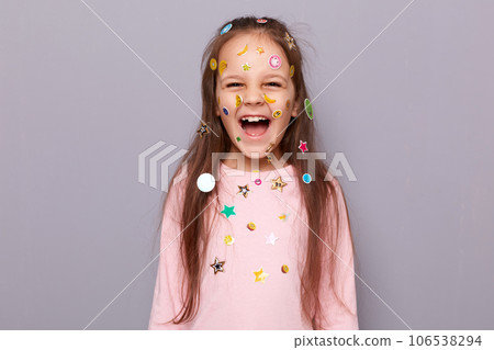 Happy joyful cheerful little girl covered colorful kid's stickers standing isolated over gray background and laughing being in good playful mood expressing positive emotions. 106538294