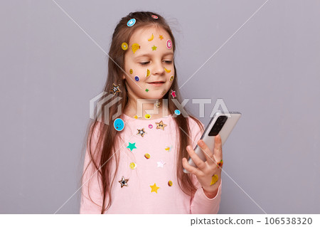 Happy cheerful little girl covered colorful kid's stickers standing isolated over gray background using smartphone for chatting with her friends in social networks. Happy cheerful little girl covered colorful kid's stickers standing isolated over gray background using smartphone for chatting with her friends in social networks. 106538320