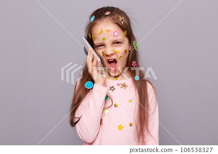 Funny crazy little girl covered colorful kid's stickers standing isolated over gray background talking on smartphone arguing with somebody grimacing making face. Funny crazy little girl covered colorful kid's stickers standing isolated over gray background talking on smartphone arguing with somebody grimacing making face. 106538327