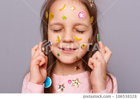 Hopeful little girl covered colorful kid's stickers standing isolated over gray background standing with closed eyes and crossed fingers making wish praying for good luck. Hopeful little girl covered colorful kid's stickers standing isolated over gray background standing with closed eyes and crossed fingers making wish praying for good luck. 106538328