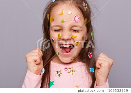 Extremely happy joyful little girl covered colorful kid's stickers standing isolated over gray background closing her eyes and clenched fists celebrating her success. 106538329