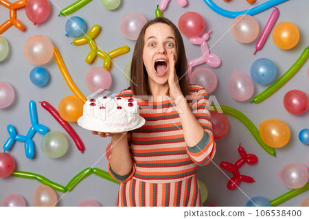 Excited woman wearing striped dress holding cake and keeps hand near mouth screaming loud making announcement inviting guests to her party standing against gray wall decorated with colorful balloons. 106538400