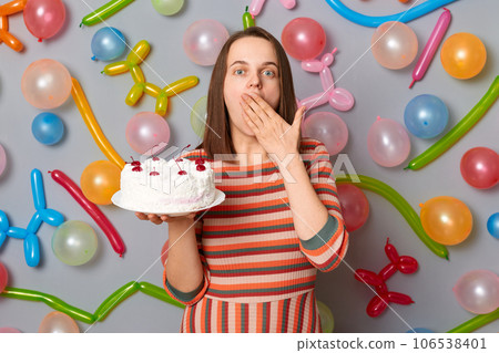 Amazed surprised woman with brown hair wearing striped dress holding cake covering mouth with hands sees shocked birthday present standing against gray wall decorated with colorful balloons. Amazed surprised woman with brown hair wearing striped dress holding cake covering mouth with hands sees shocked birthday present standing against gray wall decorated with colorful balloons. 106538401