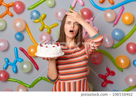 Disappointed birthday woman wearing striped dress holding cake making faceplam gesture forgot something important for her party standing against gray wall decorated with colorful balloons. 106538402