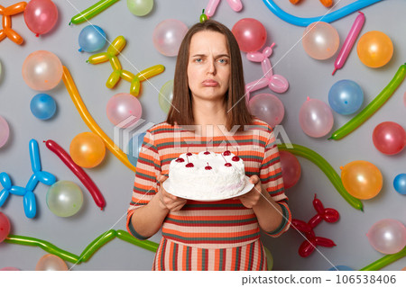 Sad disappointed woman with brown hair wearing striped dress holding cake she dislikes mixing order buying wrong dessert standing against gray wall decorated with colorful balloons. 106538406