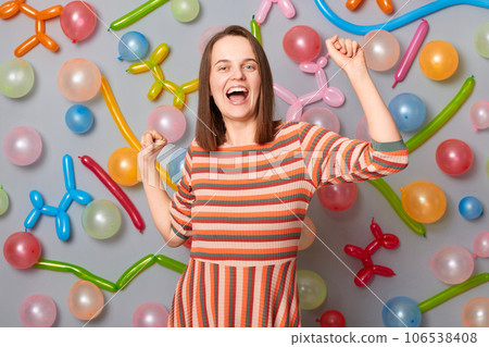 Festive happy woman wearing striped dress standing against gray wall with balloons decoration dancing with raised arms celebrating festive mood cheerful emotions. Festive happy woman wearing striped dress standing against gray wall with balloons decoration dancing with raised arms celebrating festive mood cheerful emotions. 106538408
