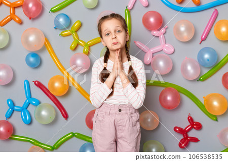Pleading child. Sad upset little girl with pigtails standing against gray wall decorated with colorful balloons keeps hands in praying gesture asking to forgive. Pleading child. Sad upset little girl with pigtails standing against gray wall decorated with colorful balloons keeps hands in praying gesture asking to forgive. 106538535