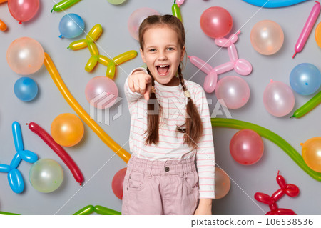 Overjoyed excited little girl with pigtails standing against gray wall with balloons decoration pointing to camera with amazement screaming celebration. Overjoyed excited little girl with pigtails standing against gray wall with balloons decoration pointing to camera with amazement screaming celebration. 106538536