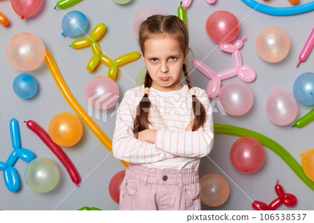 Sad offended little girl with pigtails standing against gray wall decorated with colorful balloons keeps hands folded looking at camera with pout lips. 106538537