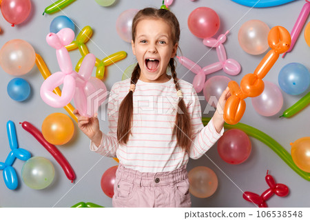 Extremely excited festive little girl with pigtails standing against gray decorated colorful wall holding balloons in hands screaming hurray celebrating birthday. Extremely excited festive little girl with pigtails standing against gray decorated colorful wall holding balloons in hands screaming hurray celebrating birthday. 106538548