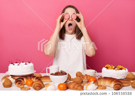 Funny crazy brown haired woman in white t shirt sitting at table with cinfectionery isolated over pink background covering eyes with cookies keeps mouth widely opened. Funny crazy brown haired woman in white t shirt sitting at table with cinfectionery isolated over pink background covering eyes with cookies keeps mouth widely opened. 106538588