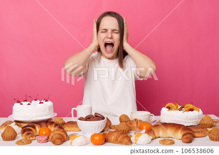 Shocked despair irritated woman wearing white T-shirt sitting at festive table with various homemade pastry keeps diet covering ears with hands screaming isolated over pink background. Shocked despair irritated woman wearing white T-shirt sitting at festive table with various homemade pastry keeps diet covering ears with hands screaming isolated over pink background. 106538606