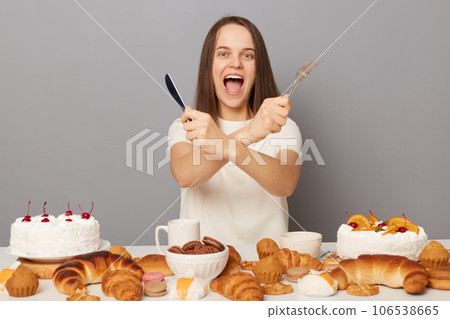 Excited woman wearing white T-shirt posing surrounded with various confectionery, isolated over gray background, crossed arms, holding knife and fork. 106538665