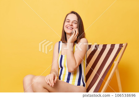 Pretty smiling woman wearing striped swimming suit sitting on deck chair isolated over yellow background talking on smart phone boasting her vacation. Pretty smiling woman wearing striped swimming suit sitting on deck chair isolated over yellow background talking on smart phone boasting her vacation. 106538696