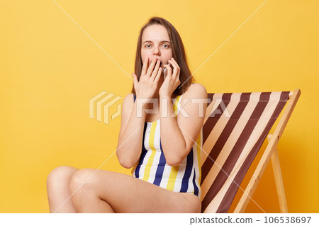 Surprised shocked woman wearing striped swimming suit sitting on deck chair talking on cell phone covering open mouth with hand isolated over yellow background. Surprised shocked woman wearing striped swimming suit sitting on deck chair talking on cell phone covering open mouth with hand isolated over yellow background. 106538697