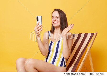 Resort blog. Smiling woman blogger wearing striped swimming suit sitting on deck chair isolated over yellow background, broadcasting livestream from vacation, having video call, saying hi. Resort blog. Smiling woman blogger wearing striped swimming suit sitting on deck chair isolated over yellow background, broadcasting livestream from vacation, having video call, saying hi. 106538702