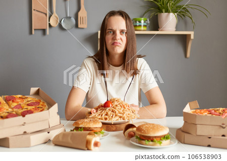 Sad unhappy woman wearing white t- shirt siting at table with fast food in kitchen looking at camera with displeased face felling overeating unhealthy nutrition. 106538903