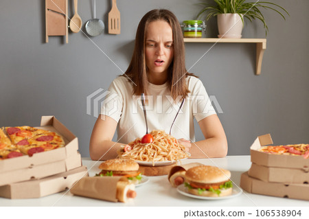Confused shocked woman wearing white t- shirt siting at table with fast food in kitchen looking at unhealthy dishes with puzzled face, ordering too much meal. 106538904