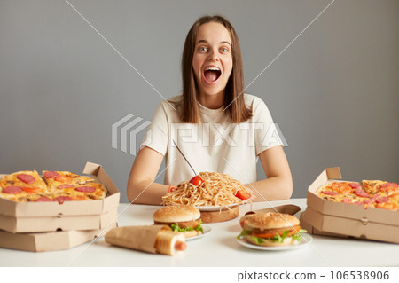Amazed excited woman with brown hair wearing white T-shirt sitting at table among unhealthy food isolated over gray background breaks diet looking at camera screaming with cheerful face. Amazed excited woman with brown hair wearing white T-shirt sitting at table among unhealthy food isolated over gray background breaks diet looking at camera screaming with cheerful face. 106538906