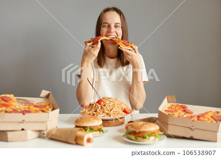 Hungry funny woman with brown hair wearing white T-shirt sitting at table among fast food isolated over gray background holding two slices of pizza eating junk food. 106538907