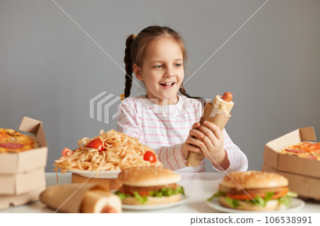 Excited charming little girl with braids sitting at table with fast food isolated over gray background holding big hot dog looking at unhealthy meal with big amazed eyes and smile. 106538991