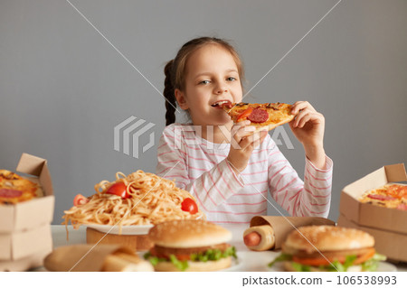 Hungry optimistic attractive little girl with braids sitting at table with junk food isolated over gray background biting big slice of pizza enjoying good appetite dinner. 106538993