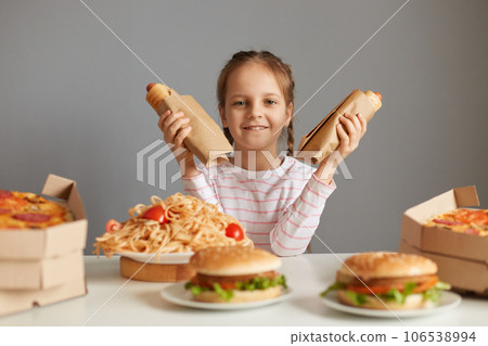 Satisfied delighted little girl with braids sitting at table with fast food isolated over gray background holding two hot dogs looking at cmaera with happy expression. 106538994