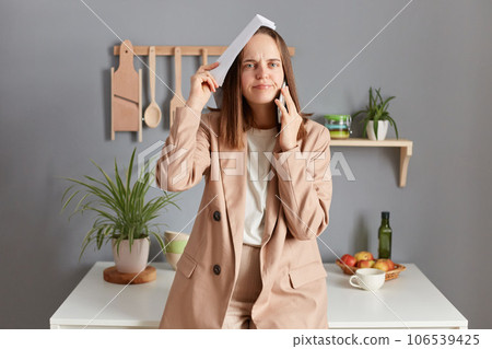 Displeased sad woman with brown hair wearing beige jacket standing in home kitchen interior having conversation with boss makes mistakes in reports expressing sorrow. 106539425