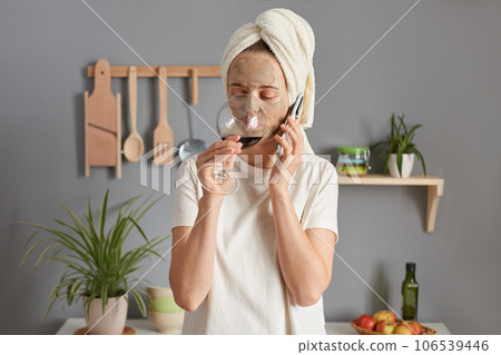 Caucasian beautiful young woman posing in kitchen with towel on head drinking wine talking on smart phone having conversation on mobile phone. Caucasian beautiful young woman posing in kitchen with towel on head drinking wine talking on smart phone having conversation on mobile phone. 106539446