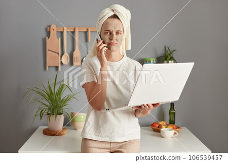 Disappointed displeased young woman posing in kitchen with towel on head working online having troubles on work holding notebook talking phone with sad expression. 106539457
