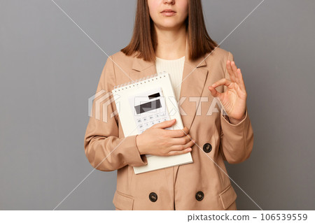 Unrecognizable woman office worker holding organizer and calculator isolated on gray background, showing ok sign, approved gesture. Unrecognizable woman office worker holding organizer and calculator isolated on gray background, showing ok sign, approved gesture. 106539559
