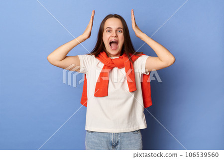 Positive cheerful surprised woman wearing white t-shirt and jumper over neck standing isolated over blue background raised arms being amazed and excited hearing great news. Positive cheerful surprised woman wearing white t-shirt and jumper over neck standing isolated over blue background raised arms being amazed and excited hearing great news. 106539560