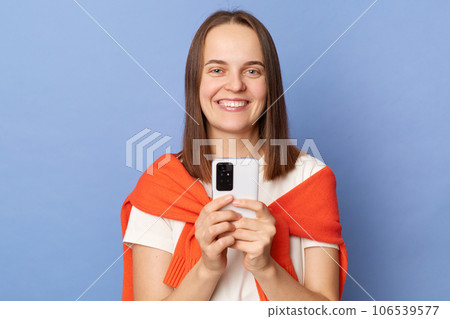 Satisfied dark haired woman wearing orange jumper over shoulders standing isolated over blue background holding mobile phone looking smiling at camera using smartphone. Satisfied dark haired woman wearing orange jumper over shoulders standing isolated over blue background holding mobile phone looking smiling at camera using smartphone. 106539577
