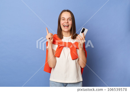 Excited inspired dark haired woman wearing orange jumper over shoulders using mobile phone raising finger pointing at copy space standing isolated over blue background. Excited inspired dark haired woman wearing orange jumper over shoulders using mobile phone raising finger pointing at copy space standing isolated over blue background. 106539578