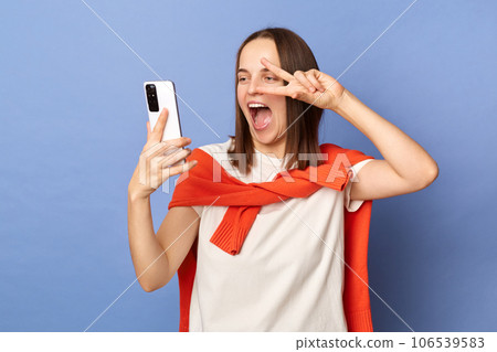 Cheerful joyful woman wearing white t-shirt and jumper over neck standing isolated over blue background standing making selfie showing victory gesture v sign. Cheerful joyful woman wearing white t-shirt and jumper over neck standing isolated over blue background standing making selfie showing victory gesture v sign. 106539583