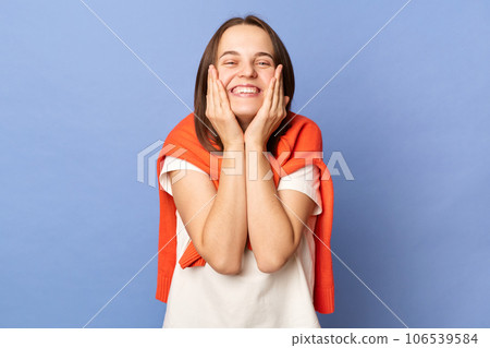 Positive extremely happy woman wearing white t-shirt and jumper over neck standing isolated over blue background keeps hands on cheeks smiling toothily. Positive extremely happy woman wearing white t-shirt and jumper over neck standing isolated over blue background keeps hands on cheeks smiling toothily. 106539584