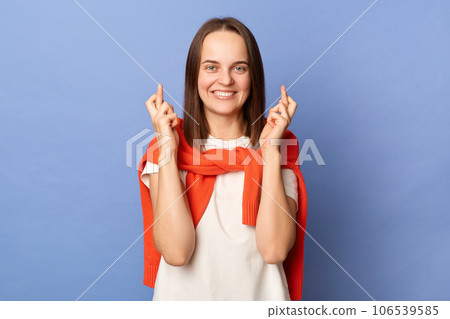 Smiling happy woman wearing white t-shirt and jumper over neck standing isolated over blue background crossing fingers dreaming for good luck looking at camera with toothy smile. 106539585